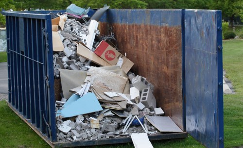 A large blue metal waste container, positioned outdoors on a paved or grassy area, is filled with mixed construction debris and general rubbish. The contents include broken concrete slabs with rough, uneven surfaces, white and grey plasterboard fragments, and assorted cardboard boxes, some of which are flattened or torn. There are also pieces of wood, plastic, and other miscellaneous discarded materials visible, with some items leaning or stacked irregularly inside the container. The container's interior is rusty brown on the back wall, contrasting with the blue exterior. The surrounding environment shows a well-maintained green lawn and trees in the background, indicating a residential or park setting. The scene suggests a rubbish removal or clearance activity, likely managed by a professional waste management service such as Commercial Waste Bromley, offering local rubbish removal solutions in Bromley and surrounding areas, including nearby postal districts in South London.