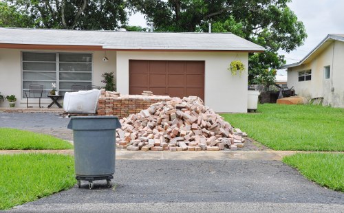 A large pile of light brown bricks and broken masonry is situated on the driveway in front of a single-story house. The house has a white exterior with a grey roof, a large window on the left with white blinds, and a brown garage door at the center. To the left of the rubble, there is a grey wheelie bin placed on the asphalt surface. The grass on either side of the driveway is well-maintained and vibrant green. In the background, surrounding trees and neighboring houses are visible, with one house partially seen on the right. The scene appears to be outdoors during daylight hours with natural lighting, and the environment suggests that this is an area where commercial waste removal services, such as those provided by Commercial Waste Bromley, might be employed for clearing construction debris or household waste in a residential setting near Bromley.