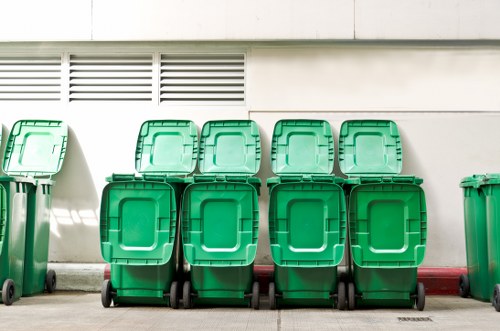 A row of eight green wheeled rubbish bins made of durable plastic with hinged lids, positioned against a light-coloured exterior wall of a building, possibly a commercial or industrial property. The bins are arranged in two tiers, with four on the ground and four stacked on top, all neatly aligned and secured with black wheels at the base. The surface beneath the bins appears to be paved, with a narrow strip of red brick or concrete edging along the wall, indicative of a typical waste collection area in Bromley. The wall behind features ventilation grilles and a horizontal band of siding, suggesting an urban or suburban setting associated with waste management services provided by Commercial Waste Bromley. The lighting is bright, indicating daylight conditions, with shadows cast on the wall, highlighting the clean and well-maintained condition of the bins and surrounding area, aligning with professional rubbish removal operations in the local area.